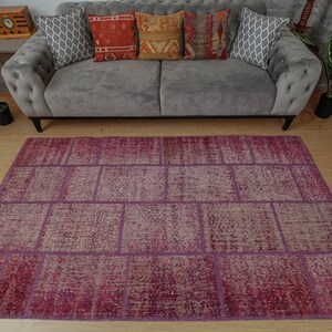 Op de afbeelding: Een patchwork tapijt met een paarse en beige kleurstelling. Het tapijt is gemaakt van verschillende stukken stof die aan elkaar zijn genaaid, waardoor een uniek en textuurrijk ontwerp ontstaat.