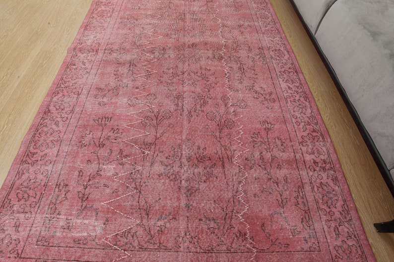 May include: A rectangular rug in shades of pink and faded black, with a distressed appearance. It features a central floral design with a zigzag pattern and a floral border. The rug is placed on a light-coloured wooden floor.