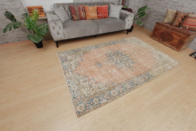 Puede incluir: Una alfombra vintage con un centro melocot&oacute;n descolorido y un borde azul y crema. La alfombra tiene un patr&oacute;n floral y est&aacute; colocada sobre un suelo de madera clara.