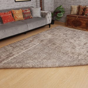 May include: A large, rectangular, distressed-look rug with a faded, muted color palette of brown and beige. The rug is partially rolled up, revealing its underside. The rug is placed on a light-colored wooden floor, with a gray sofa and decorative pillows in the background.