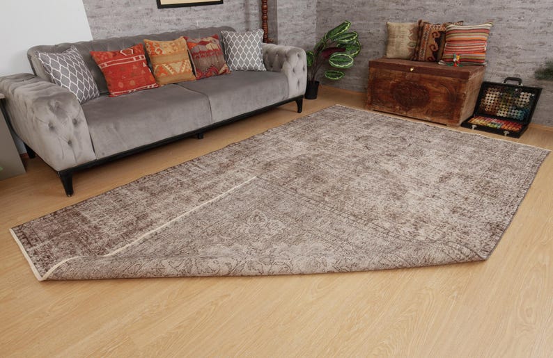 May include: A large, rectangular, distressed-look rug with a faded, muted color palette of brown and beige. The rug is partially rolled up, revealing its underside. The rug is placed on a light-colored wooden floor, with a gray sofa and decorative pillows in the background.