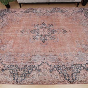 May include: A large rectangular area rug featuring a faded salmon pink base color with intricate dark blue and grey floral patterns. The design includes a central medallion and a wide, ornate border with detailed botanical motifs throughout.