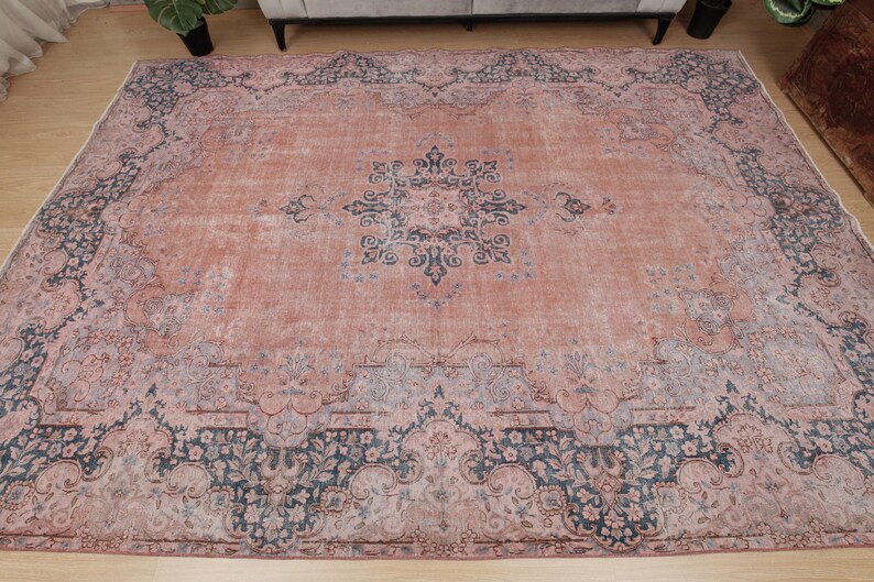 May include: A large rectangular area rug featuring a faded salmon pink base color with intricate dark blue and grey floral patterns. The design includes a central medallion and a wide, ornate border with detailed botanical motifs throughout.