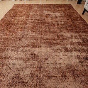 May include: A large rectangular area rug featuring a distressed, weathered appearance in shades of deep brown, copper, and faded tan. The surface displays a complex, mottled texture with subtle linear patterns and varying color intensity throughout.