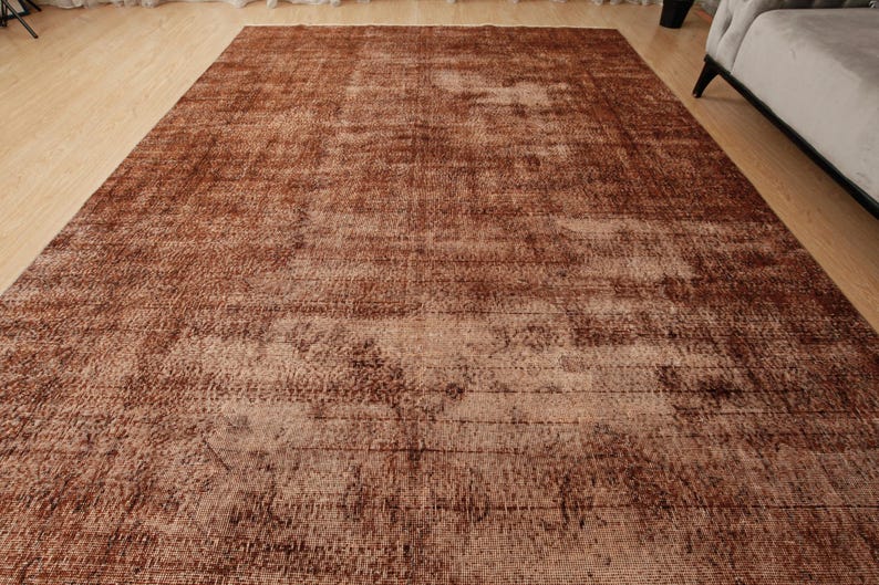 May include: A large rectangular area rug featuring a distressed, weathered appearance in shades of deep brown, copper, and faded tan. The surface displays a complex, mottled texture with subtle linear patterns and varying color intensity throughout.