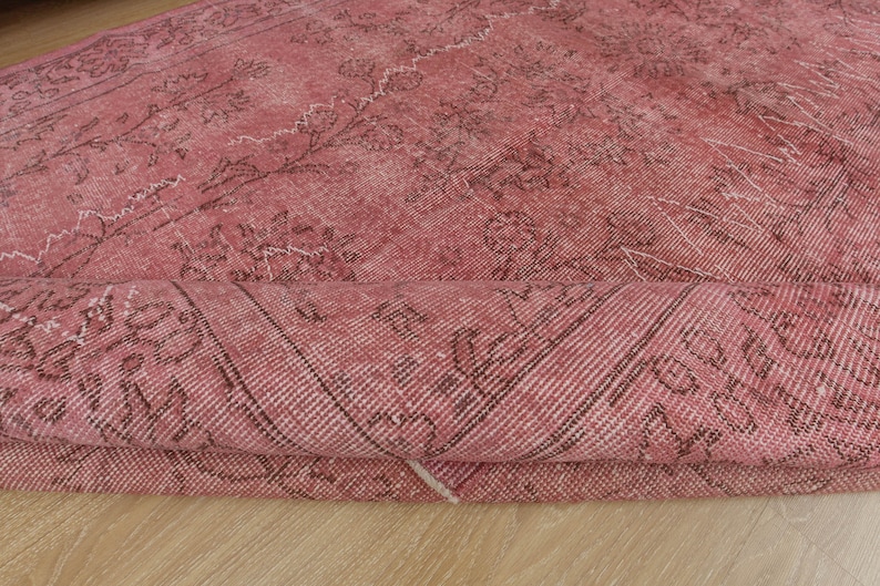 May include: A rolled-up, distressed pink rug with a faded floral pattern. The rug features dark brown and white accents, creating a vintage aesthetic. The rug is made of a woven material.