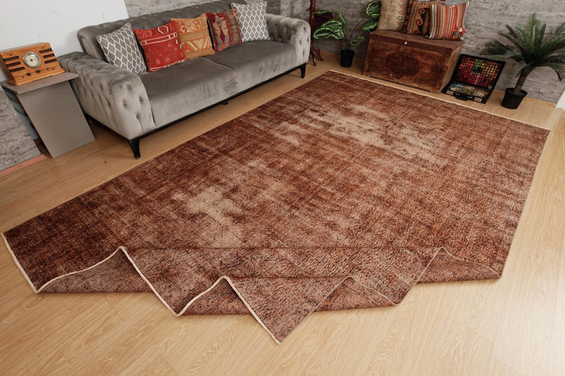 May include: A large rectangular area rug featuring a distressed, weathered texture in shades of deep brown and copper. The rug has a faded, mottled appearance with a visible folded corner revealing a light-colored edge binding on the underside.