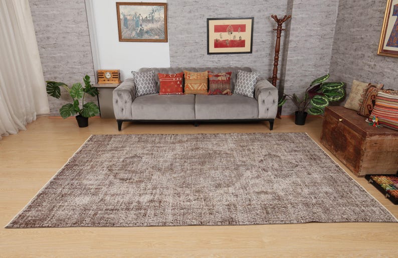 May include: A large, rectangular, distressed-look rug in shades of brown and beige dominates the foreground. A gray sofa with decorative pillows sits against a textured wall, with artwork and plants adding to the room's decor.