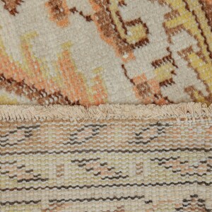 May include: Close-up of a beige rug with a detailed pattern in shades of orange, yellow, and brown. The rug has a fringed edge and a woven texture, showcasing a traditional design.