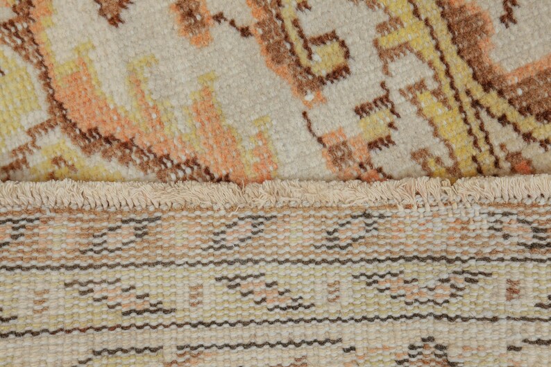 May include: Close-up of a beige rug with a detailed pattern in shades of orange, yellow, and brown. The rug has a fringed edge and a woven texture, showcasing a traditional design.