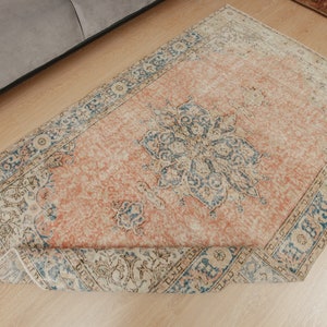 Puede incluir: Una alfombra vintage con fondo color melocot&oacute;n y un patr&oacute;n floral azul y blanco. La alfombra tiene un borde con flecos y est&aacute; enrollada.