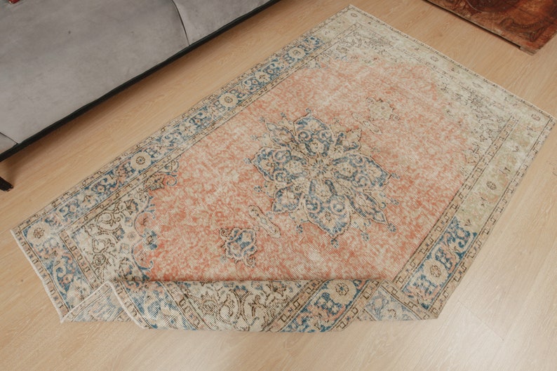 Puede incluir: Una alfombra vintage con fondo color melocot&oacute;n y un patr&oacute;n floral azul y blanco. La alfombra tiene un borde con flecos y est&aacute; enrollada.