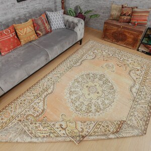 May include: An overhead shot of a large, rectangular, peach-colored rug with an intricate floral pattern. The rug is partially rolled up, revealing its detailed border. A gray sofa with decorative pillows and a wooden chest are in the background.