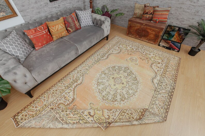 May include: An overhead shot of a large, rectangular, peach-colored rug with an intricate floral pattern. The rug is partially rolled up, revealing its detailed border. A gray sofa with decorative pillows and a wooden chest are in the background.