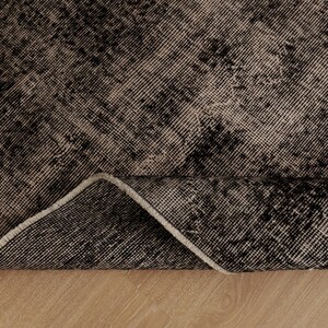 May include: A rectangular area rug featuring a distressed, mottled pattern in shades of black and off-white. The rug is folded at one corner, revealing the woven texture of the fabric and the underside edge with a light-coloured binding.