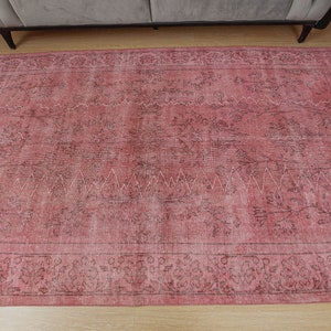 May include: A large, rectangular rug in shades of pink and brown with a distressed appearance. It has an ornate floral border and a central design with a zigzag pattern. The rug is on a light-coloured wooden floor.