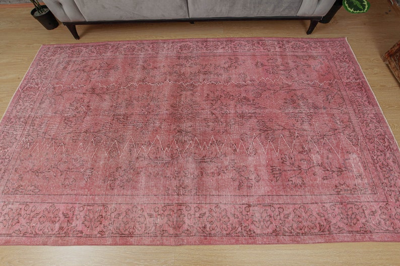 May include: A large, rectangular rug in shades of pink and brown with a distressed appearance. It has an ornate floral border and a central design with a zigzag pattern. The rug is on a light-coloured wooden floor.