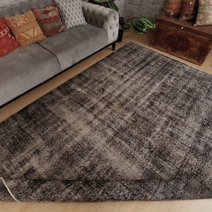 May include: A large rectangular area rug featuring a distressed, mottled pattern in shades of charcoal grey, black, and faded white. The rug has a low-pile texture with a worn, vintage-inspired appearance and a finished edge binding.