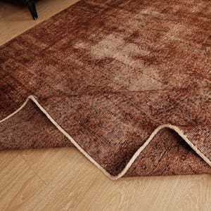 May include: A rectangular area rug featuring a distressed, faded pattern in shades of deep brown, rust, and tan. The rug has a folded corner revealing the underside, showcasing a woven texture and a light-colored, finished edge binding.