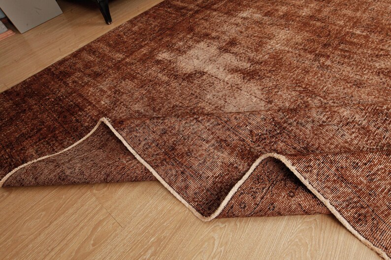 May include: A rectangular area rug featuring a distressed, faded pattern in shades of deep brown, rust, and tan. The rug has a folded corner revealing the underside, showcasing a woven texture and a light-colored, finished edge binding.
