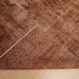 May include: A rectangular area rug in shades of rust and brown with a distressed, weathered texture. One corner of the rug is folded over to reveal the underside, which displays a darker, intricate, and symmetrical floral pattern in black.