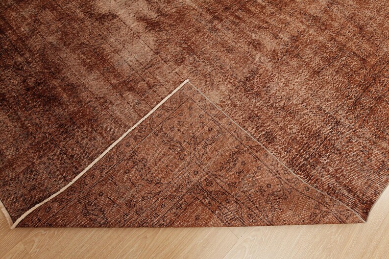 May include: A rectangular area rug in shades of rust and brown with a distressed, weathered texture. One corner of the rug is folded over to reveal the underside, which displays a darker, intricate, and symmetrical floral pattern in black.