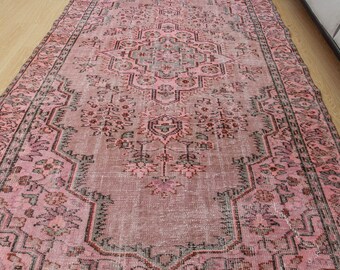 May include: A rectangular, distressed pink rug with an intricate, symmetrical floral and geometric pattern. The rug features a central medallion design and a border with repeating motifs. The color palette includes shades of pink, gray, and black.
