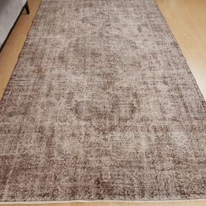 May include: A rectangular, distressed-look rug with a faded, vintage appearance. The rug features a central medallion design and a border, with a color palette of brown and cream. The rug is placed on a hardwood floor.