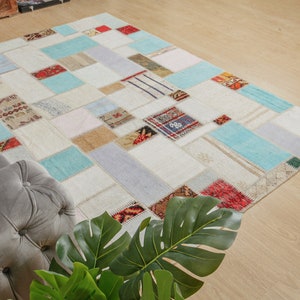 Puede incluir: Una alfombra de patchwork con fondo blanco y parches de varios colores, incluyendo azul, rojo y beige. La alfombra está hecha de diferentes telas y texturas, creando un aspecto único y ecléctico.