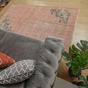 May include: A gray sofa with a gray and white patterned pillow and a red and orange patterned pillow. The sofa is sitting on a pink and white floral patterned rug. A green plant is in the background.