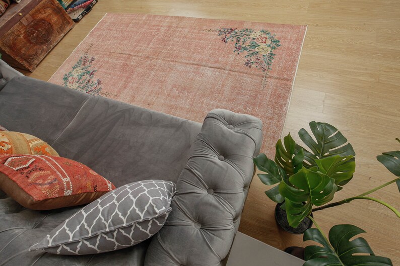 May include: A gray sofa with a gray and white patterned pillow and a red and orange patterned pillow. The sofa is sitting on a pink and white floral patterned rug. A green plant is in the background.