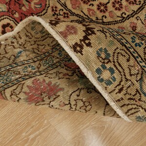 Pu&ograve; includere: Primo piano di una sezione di un tappeto vintage con un bordo piegato. Il tappeto presenta uno sfondo beige con intricati motivi floreali in rosso, blu e marrone. La trama e il design suggeriscono che si tratti di un oggetto d'antiquariato o di una riproduzione di alta qualit&agrave;.