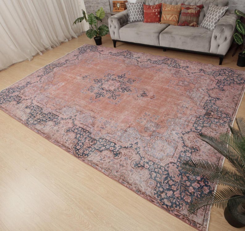 May include: A large rectangular area rug featuring a faded terracotta orange center with a dark blue floral medallion. The border displays intricate dark blue and muted grey floral patterns against a light terracotta background, creating a distressed look.
