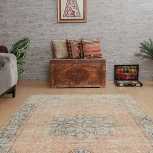 Puede incluir: Una alfombra vintage con fondo de color melocot&oacute;n claro y un patr&oacute;n floral azul y blanco. La alfombra est&aacute; colocada sobre un suelo de madera delante de un cofre de madera con almohadas en la parte superior.