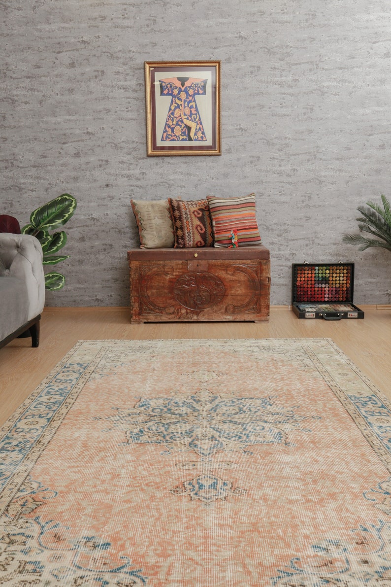Puede incluir: Una alfombra vintage con fondo de color melocot&oacute;n claro y un patr&oacute;n floral azul y blanco. La alfombra est&aacute; colocada sobre un suelo de madera delante de un cofre de madera con almohadas en la parte superior.
