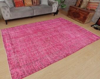 May include: A large, rectangular, distressed pink rug with a faded, vintage appearance. The rug has a textured surface with variations in colour, creating a worn look. The rug is placed on a light-coloured wooden floor, with a grey sofa and decorative cushions in the background.