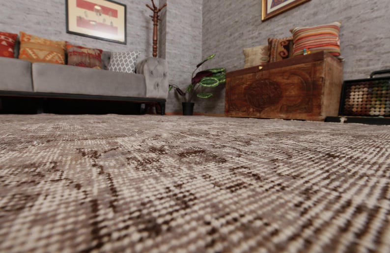 May include: A low-angle shot of a patterned rug in a room with a gray sofa, wooden chest, and framed artwork. The rug has a beige and brown design, with a textured appearance. The room has a neutral color palette, creating a cozy atmosphere.