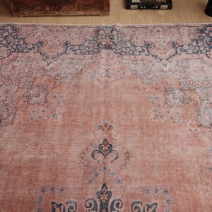 May include: A large rectangular area rug featuring a faded salmon pink field with intricate floral and vine motifs in muted blue and grey tones. The design includes a prominent central medallion and a wide decorative border with ornate patterns.
