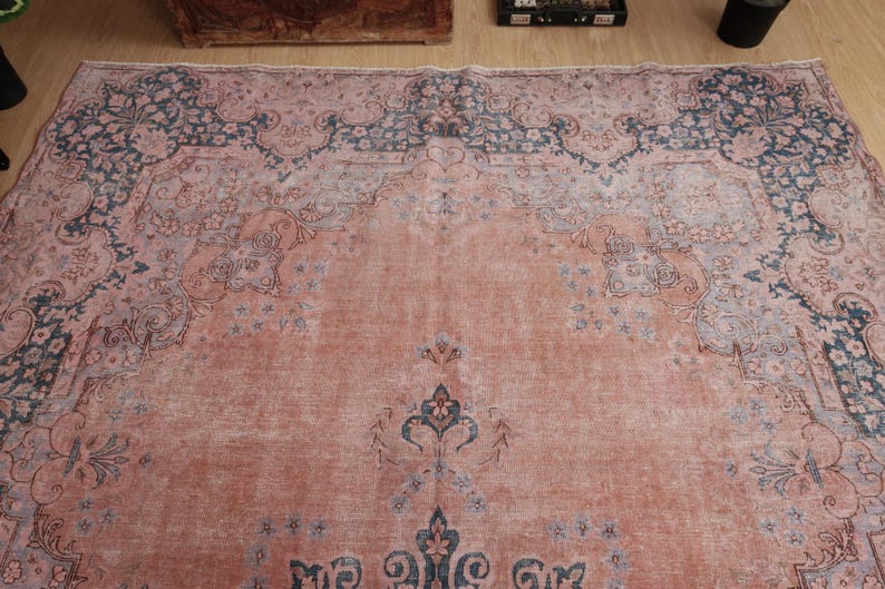 May include: A large rectangular area rug featuring a faded salmon pink field with intricate floral and vine motifs in muted blue and grey tones. The design includes a prominent central medallion and a wide decorative border with ornate patterns.