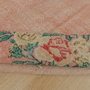 May include: Close-up of a pink rug with a floral pattern. The rug is folded in half, showing the underside of the rug. The floral pattern is in shades of green, yellow, and red.