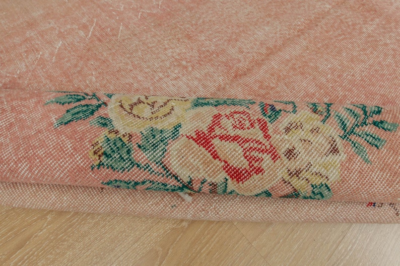 May include: Close-up of a pink rug with a floral pattern. The rug is folded in half, showing the underside of the rug. The floral pattern is in shades of green, yellow, and red.