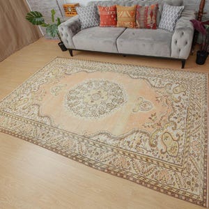 May include: A large, rectangular, vintage-style rug with an intricate floral pattern in shades of peach, cream, and brown. The rug features a central medallion design and a detailed border. The rug is placed on a light-colored wooden floor.