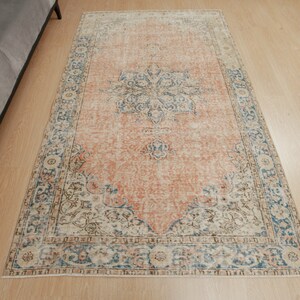 Puede incluir: Una alfombra de estilo vintage con un fondo melocot&oacute;n descolorido y un patr&oacute;n floral en tonos azules, marrones y crema. La alfombra tiene un aspecto desgastado y un borde con flecos.