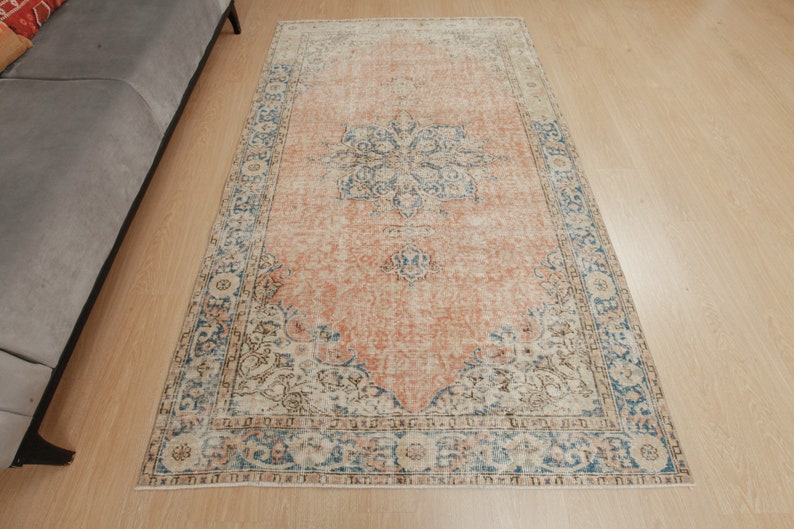 Puede incluir: Una alfombra de estilo vintage con un fondo melocot&oacute;n descolorido y un patr&oacute;n floral en tonos azules, marrones y crema. La alfombra tiene un aspecto desgastado y un borde con flecos.