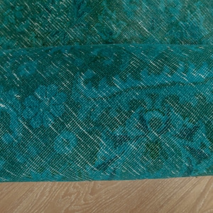 May include: Close-up of a teal blue rug with a faded floral pattern. The rug is rolled up and the edge is visible.