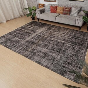 May include: A large rectangular area rug featuring a distressed, faded pattern in shades of charcoal grey, black, and white. The rug has a low-pile texture with a weathered appearance, covering a significant portion of the light-coloured wooden floor.