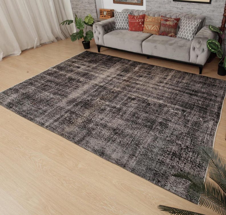 May include: A large rectangular area rug featuring a distressed, faded pattern in shades of charcoal grey, black, and white. The rug has a low-pile texture with a weathered appearance, covering a significant portion of the light-coloured wooden floor.