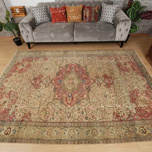 Pu&ograve; includere: Un grande tappeto rettangolare con un intricato motivo floreale nei toni del beige, rosso e blu. Il tappeto presenta un design a medaglione centrale e un bordo dettagliato. Il tappeto &egrave; posato su un pavimento in legno, con un divano grigio e cuscini decorativi sullo sfondo.