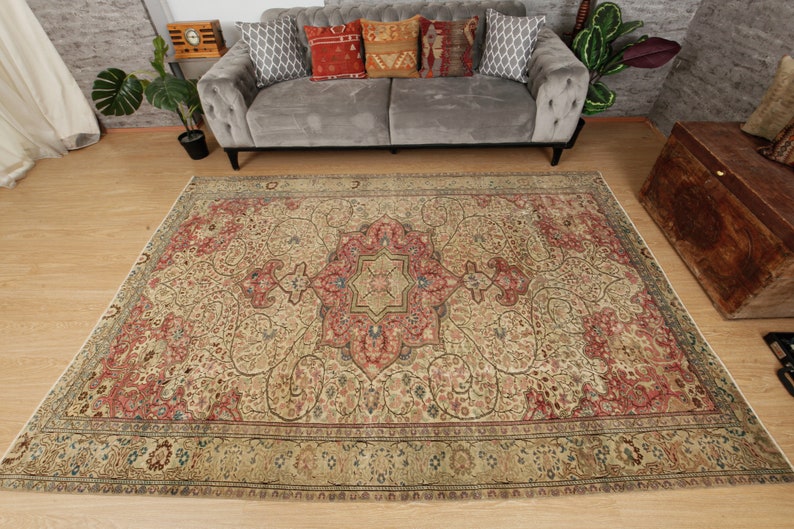 Pu&ograve; includere: Un grande tappeto rettangolare con un intricato motivo floreale nei toni del beige, rosso e blu. Il tappeto presenta un design a medaglione centrale e un bordo dettagliato. Il tappeto &egrave; posato su un pavimento in legno, con un divano grigio e cuscini decorativi sullo sfondo.