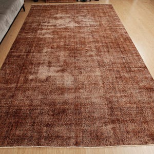 May include: A rectangular area rug featuring a deep brown and terracotta color palette with a distressed, faded texture. The rug displays a subtle, worn-in pattern across its surface, with short fringe detailing visible along the short edges.